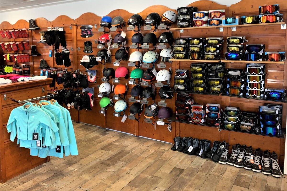 A ski/snowboard shop interior, packed with colorful helmets, goggles, boots, and winter apparel displayed on wooden slat walls and shelves.