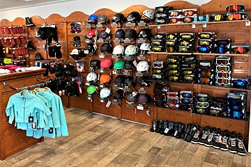 A ski/snowboard shop interior, packed with colorful helmets, goggles, boots, and winter apparel displayed on wooden slat walls and shelves.