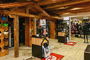 A rustic ski/snowboard shop interior with exposed wooden beams, displaying helmets, skis, boots, and winter clothing. Two checkout counters are visible.