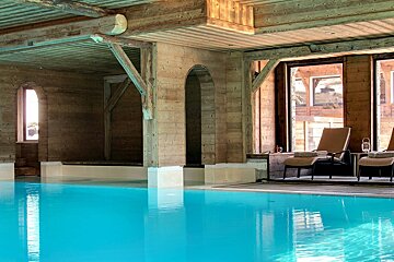 A serene indoor swimming pool with clear blue water in a rustic wooden chalet. Lounge chairs sit by large windows offering an outside view.