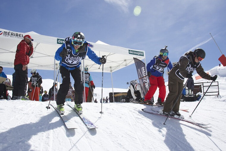 A group of skiers are going down a snow covered slope under a tent that says meribel