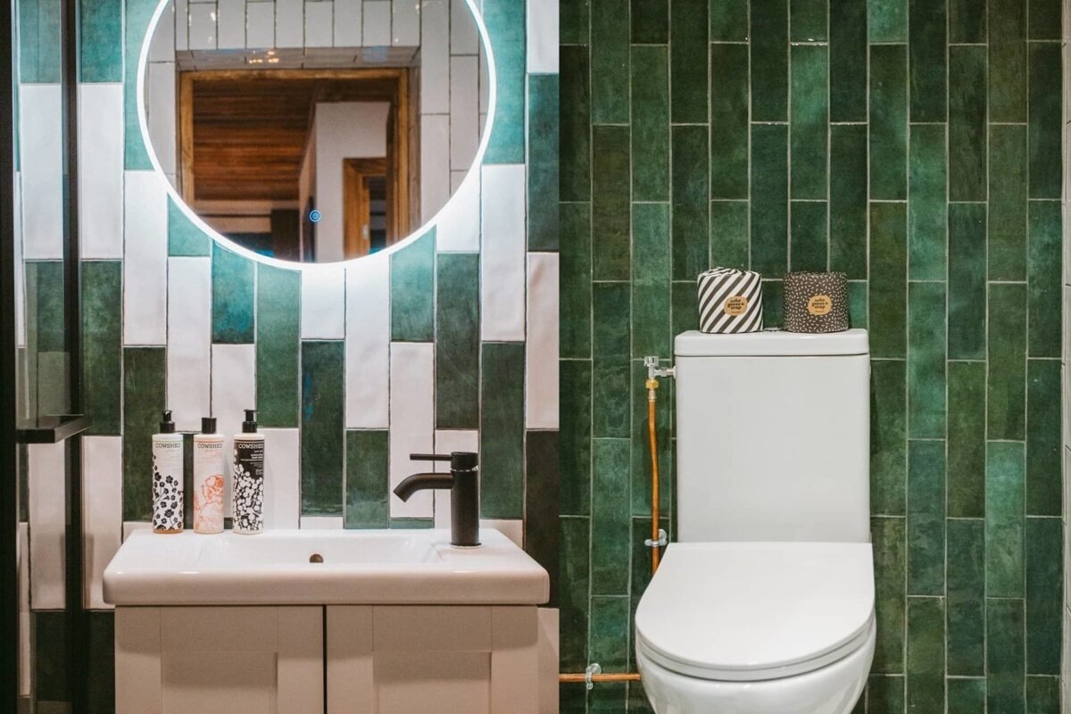 Vibrant green and white tiled bathroom featuring an illuminated mirror, a stylish sink, and a toilet with unique patterned paper.