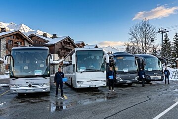 Four buses with staff are parked in a snowy mountain resort, featuring wooden chalets, snow-capped peaks, and a ski lift under a clear sky.