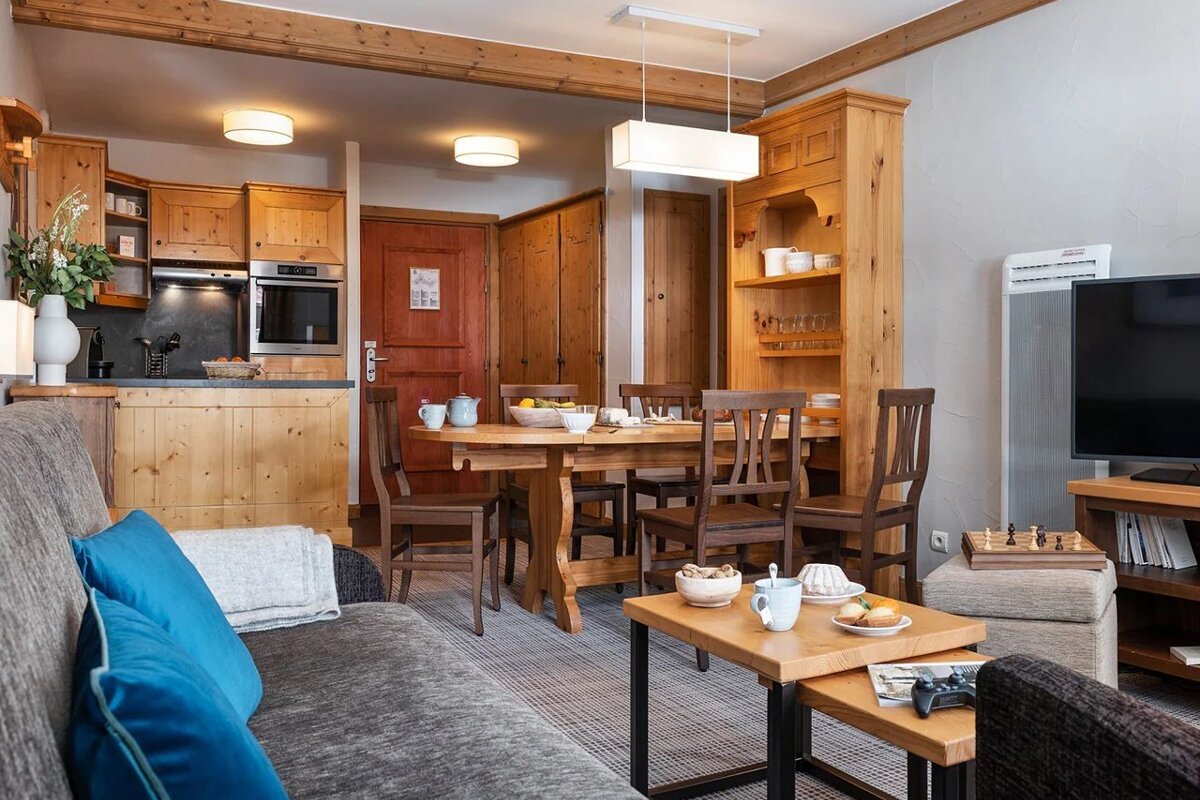 Cozy alpine-style apartment interior with wooden furniture, a dining area, a sofa in the foreground, and an open kitchen.