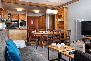 Cozy alpine-style apartment interior with wooden furniture, a dining area, a sofa in the foreground, and an open kitchen.