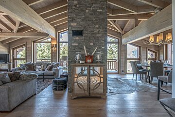 A spacious, luxurious chalet interior with exposed wood beams, a stone column, comfy sofas, a dining area, and large windows framing mountain views.
