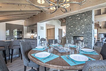 An elegant chalet dining room featuring a set table, adjacent modern kitchen, stone fireplace, and rustic ceiling beams with an antler chandelier.