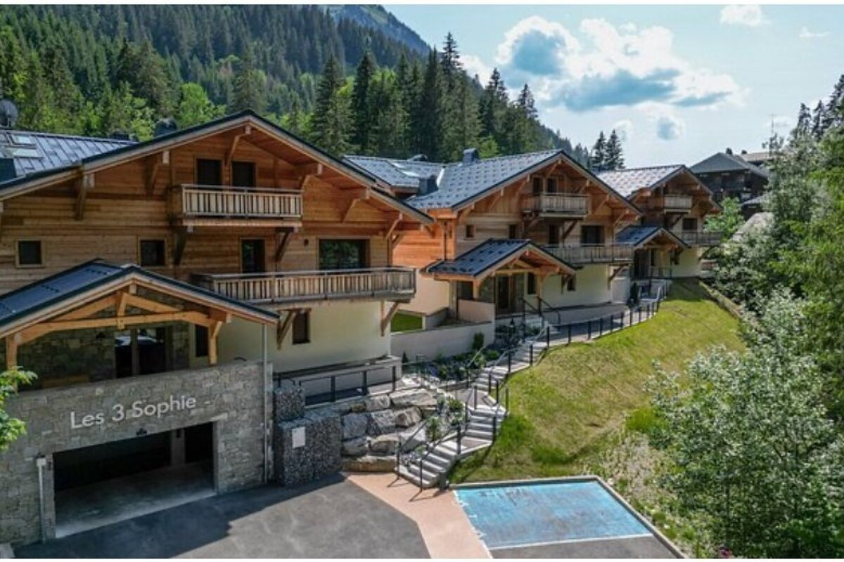 Multiple tiered wooden chalets with balconies nestled on a green mountain hillside, backed by a dense forest, named 