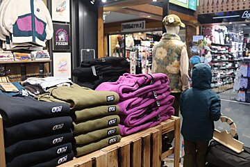 Inside a clothing store, colorful stacks of Picture brand apparel (fleeces, sweatpants) are displayed. A child in a hooded jacket stands near a mannequin.