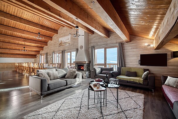 A spacious, inviting wooden chalet interior with a cozy living area, fireplace, dining hall, and stunning snowy mountain views.