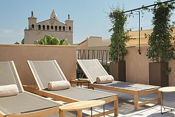 Sunny rooftop terrace with lounge chairs, rolled towels, and potted greenery, set against an old stone church and blue sky.
