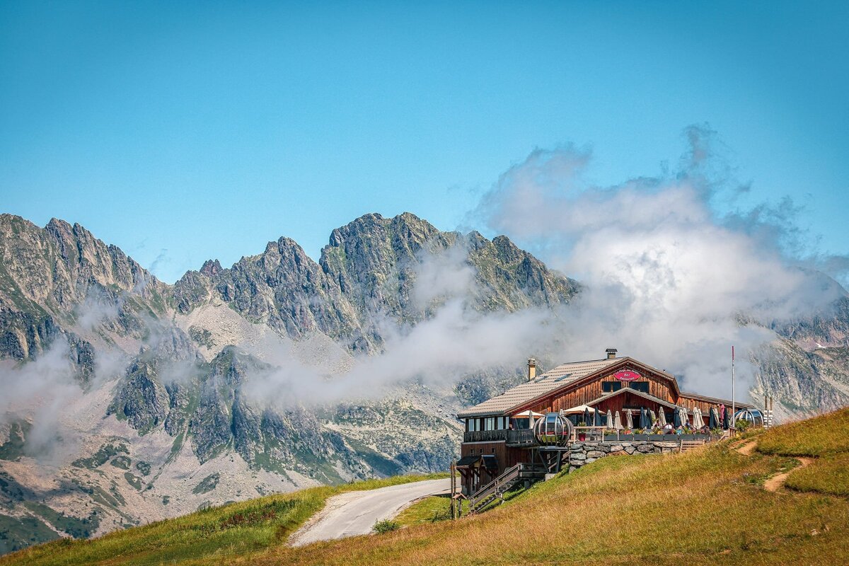 A rustic mountain lodge sits on a grassy slope, backed by rugged, cloud-shrouded peaks under a clear blue sky.