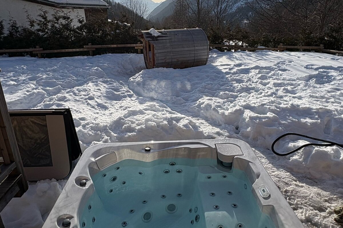 A bubbling hot tub and a barrel sauna sit amidst deep snow on a sunny winter day, with mountains in the background.