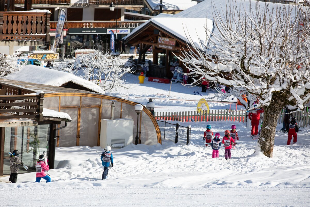 Kids in colorful ski suits play in the snow at a sunny ski resort with buildings and a ski school visible.