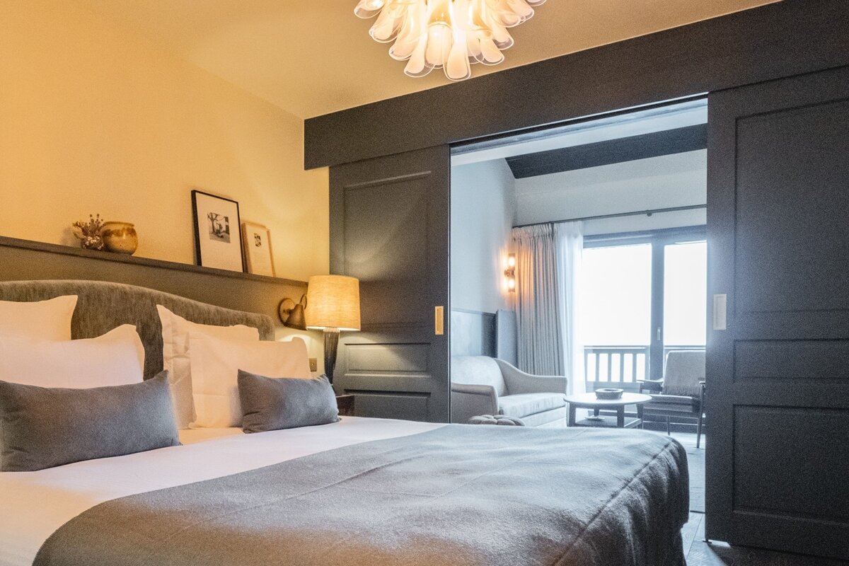 A modern, cozy bedroom with a large bed, warm lighting, and a dark sliding door opening to a bright sitting area with a balcony view.