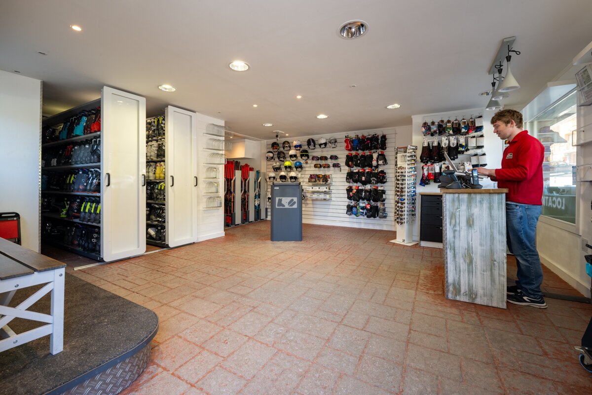 A brightly lit ski shop interior with a man at the counter. Displays show ski boots, helmets, gloves, and skis ready for rental or purchase.