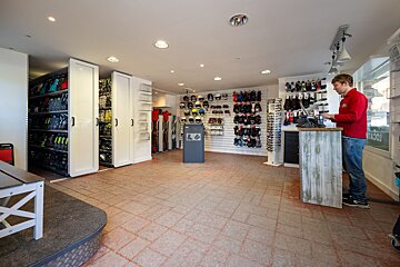 A brightly lit ski shop interior with a man at the counter. Displays show ski boots, helmets, gloves, and skis ready for rental or purchase.