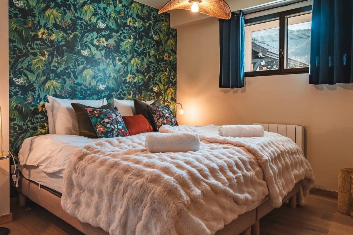 A cozy bedroom with bold jungle print wallpaper, a large bed with fluffy blanket, and a window revealing a mountain view. Warm lighting.