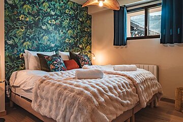 A cozy bedroom with bold jungle print wallpaper, a large bed with fluffy blanket, and a window revealing a mountain view. Warm lighting.
