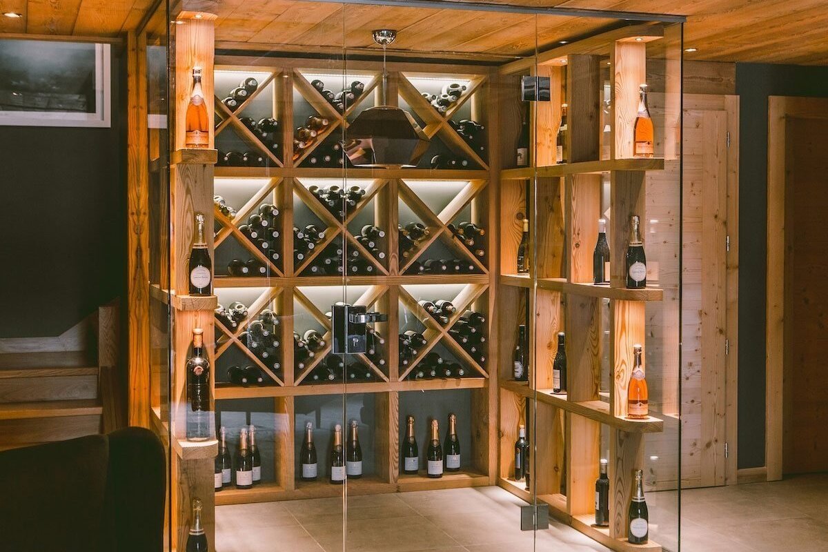 A modern, wood-paneled wine cellar with a glass enclosure showcasing numerous wine and champagne bottles in illuminated X-shaped racks.