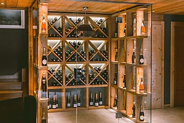 A modern, wood-paneled wine cellar with a glass enclosure showcasing numerous wine and champagne bottles in illuminated X-shaped racks.