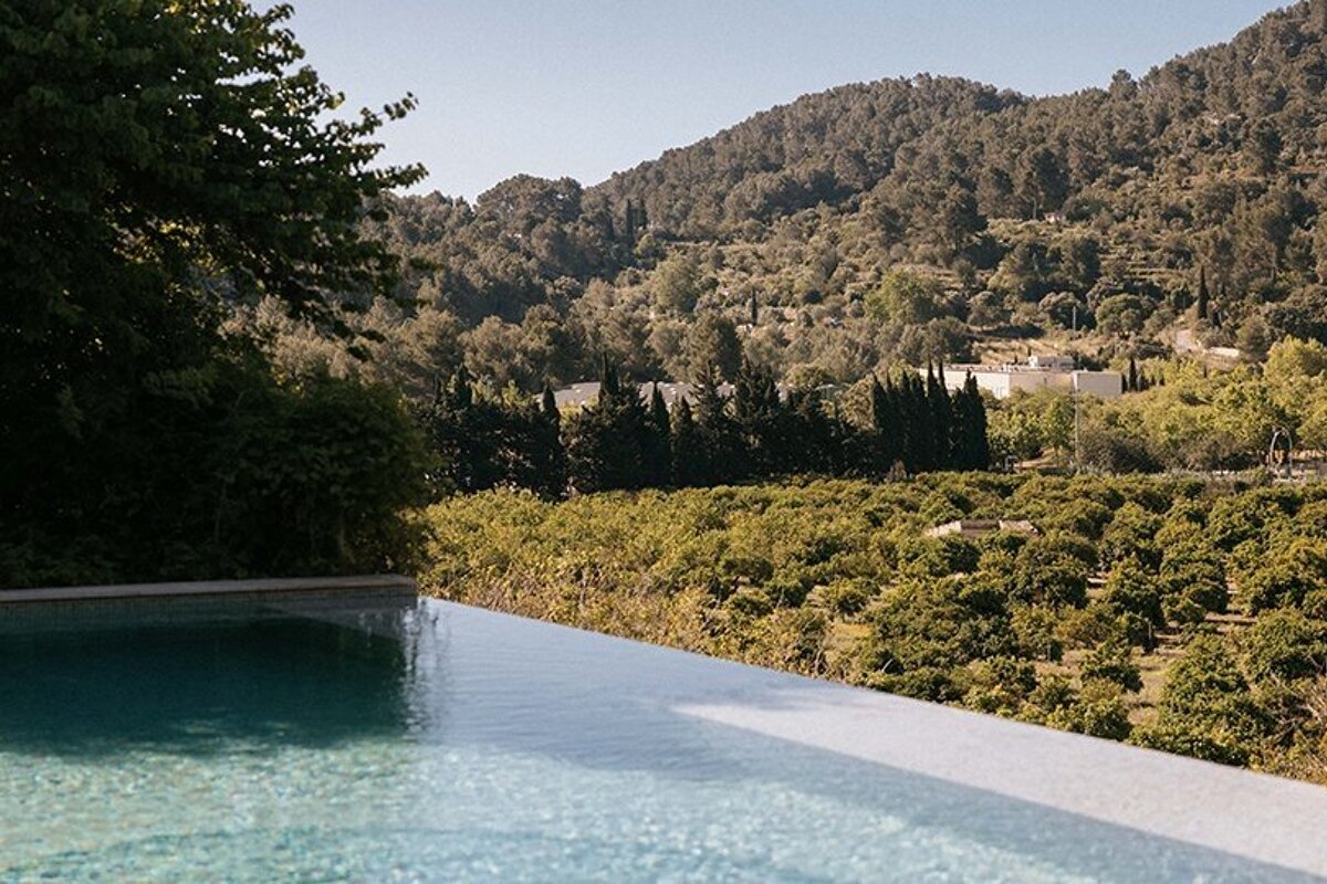 An infinity pool overlooks a lush green valley with groves, distant wooded hills, and white buildings under a clear sky.