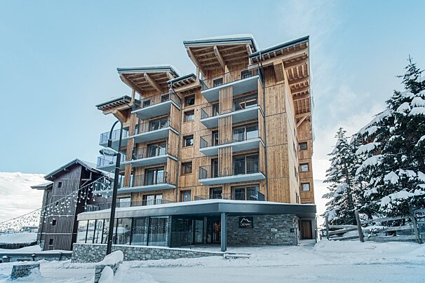 A modern, multi-story wooden building with balconies stands in a snowy mountain setting, flanked by snow-covered pine trees.