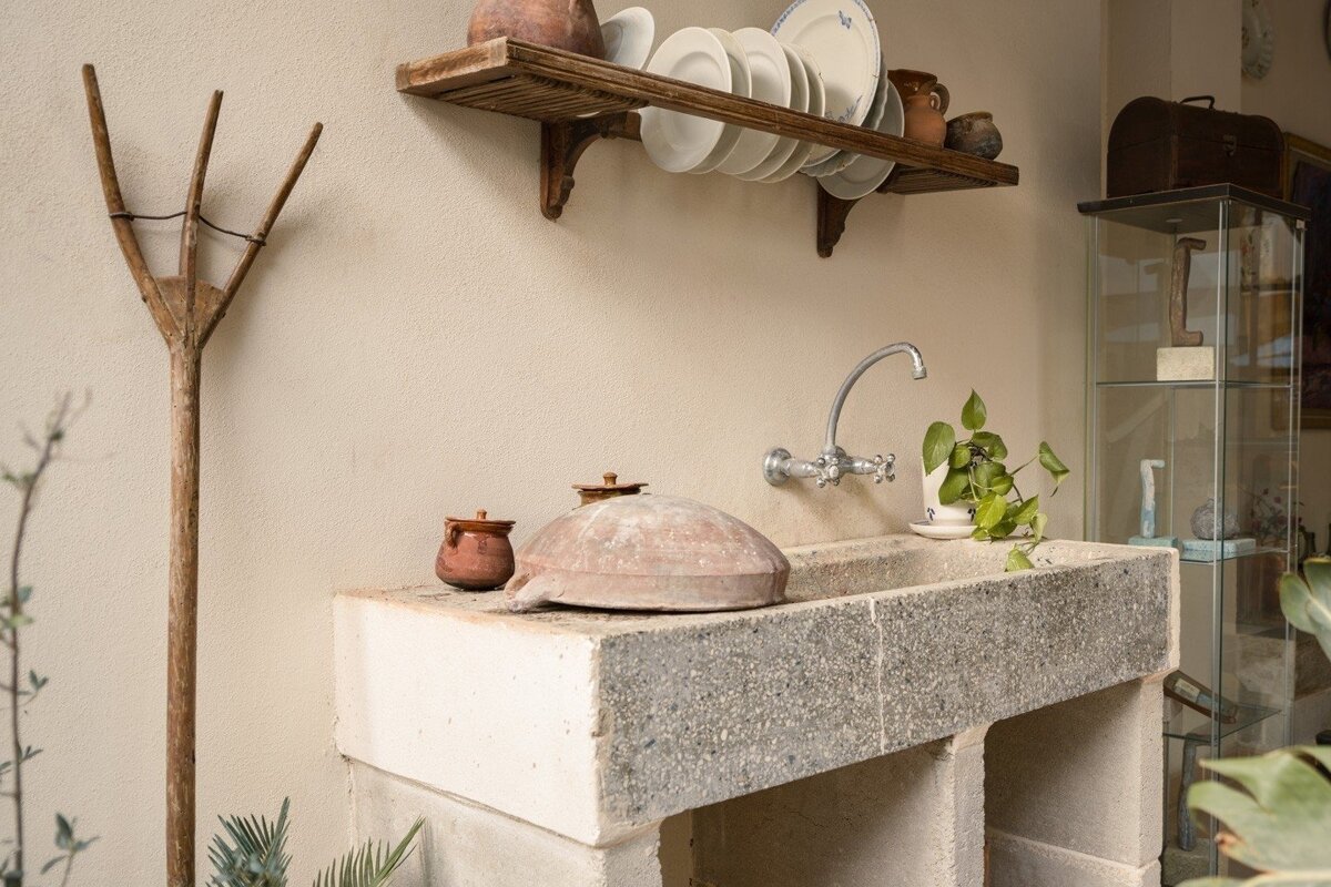 A rustic utility area featuring a stone sink, metal faucet, wooden dish rack with plates, and an old wooden farm tool.