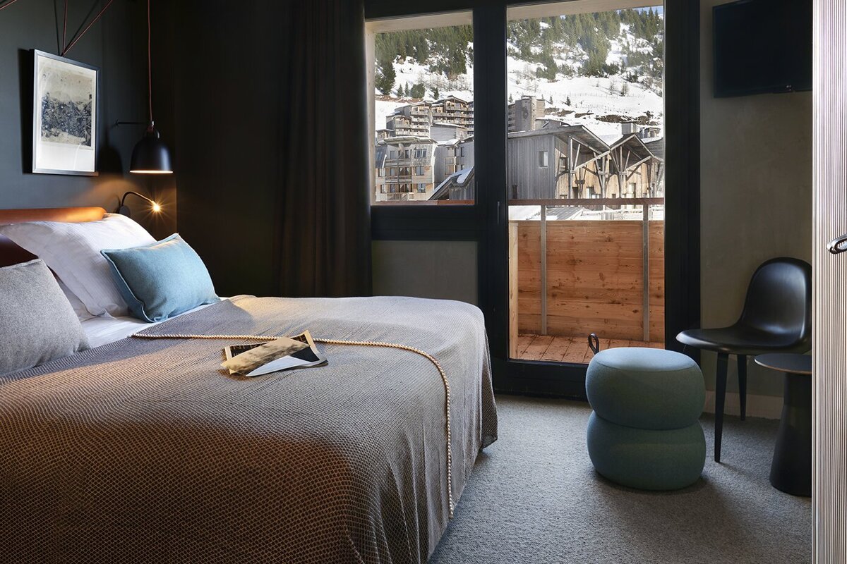A modern, cozy bedroom with dark walls and a comfortable bed, featuring a large window and balcony overlooking a snowy mountain village.