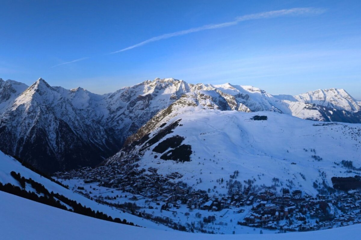 A panoramic view of snow-covered mountains with a village nestled in the valley below, under a clear blue sky with a faint contrail.