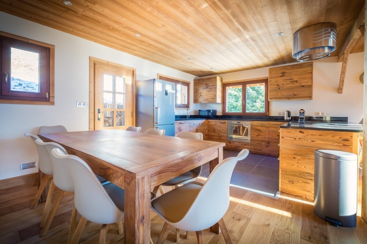 A bright, rustic-modern kitchen and dining area with extensive warm wood finishes, sleek appliances, and a large dining table with white chairs.