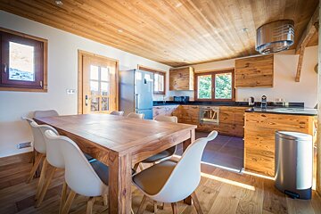 A bright, rustic-modern kitchen and dining area with extensive warm wood finishes, sleek appliances, and a large dining table with white chairs.