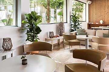 A bright, contemporary indoor lounge area with large windows, lush green plants, comfortable beige seating, and natural wood accents.