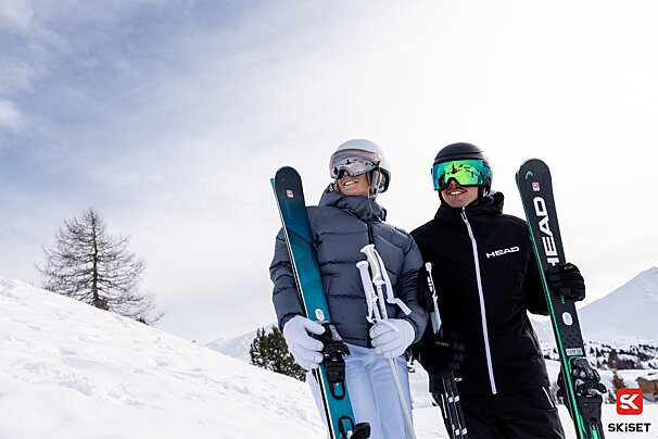 A man and a woman are holding skis and wearing head jackets