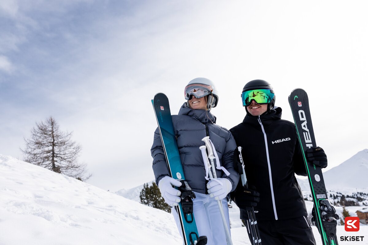Two smiling skiers, fully geared in winter attire, stand on a snowy mountain holding their skis and poles, enjoying a bright outdoor day.