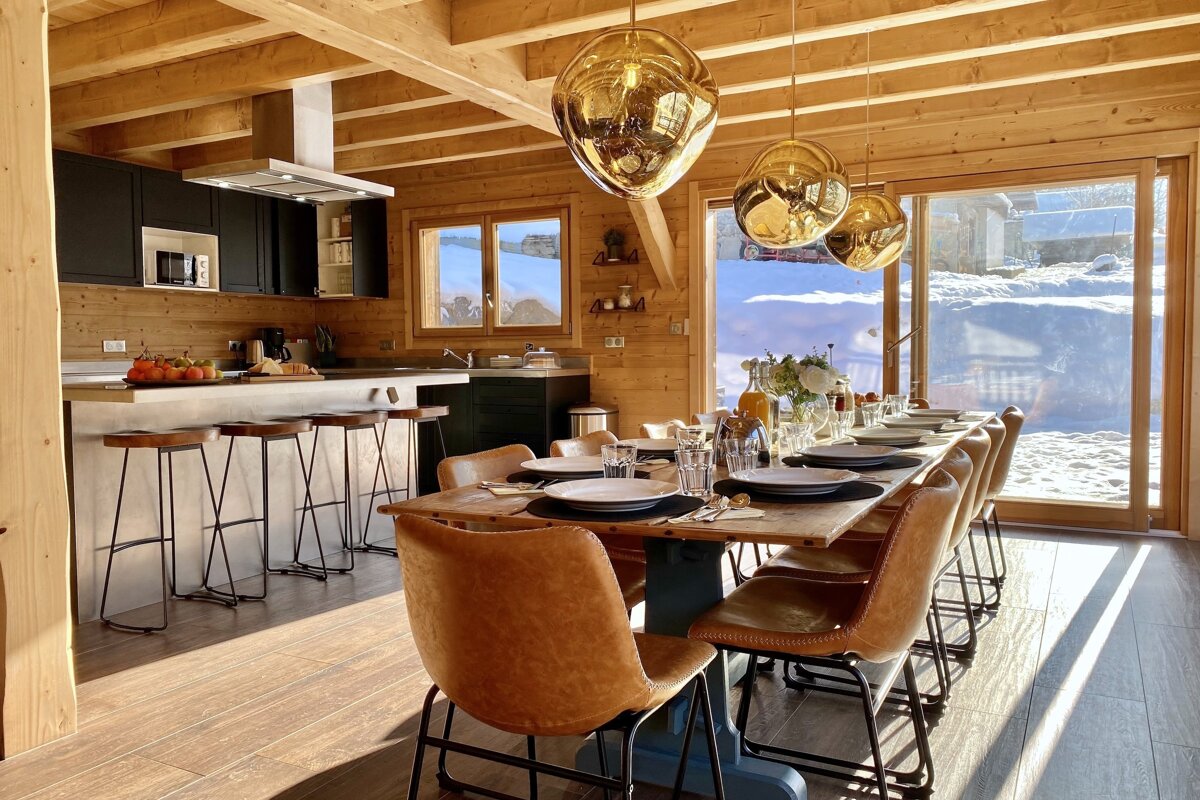 Cozy wooden cabin with a modern kitchen and dining table, bathed in sunlight, offering panoramic views of a bright, snow-covered mountain scene.