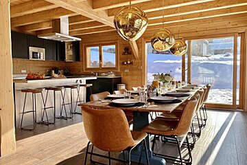 Cozy wooden cabin with a modern kitchen and dining table, bathed in sunlight, offering panoramic views of a bright, snow-covered mountain scene.