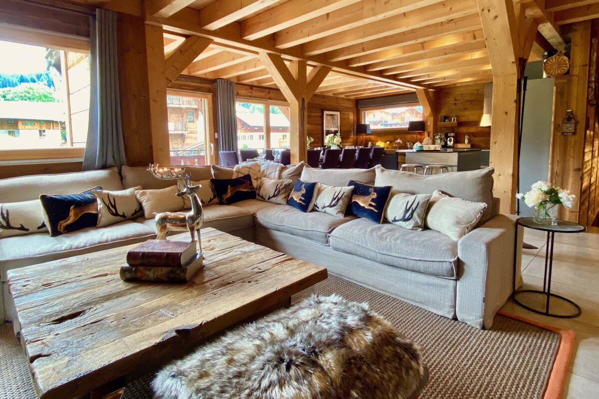 A cozy, rustic open-plan living room featuring exposed wooden beams, a large sofa with deer motif pillows, a wooden coffee table, and outdoor views.