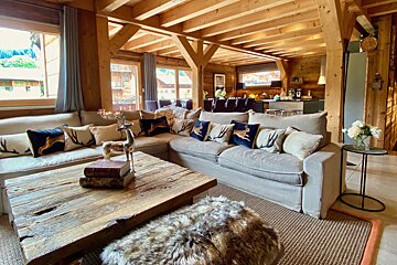 A cozy, rustic open-plan living room featuring exposed wooden beams, a large sofa with deer motif pillows, a wooden coffee table, and outdoor views.