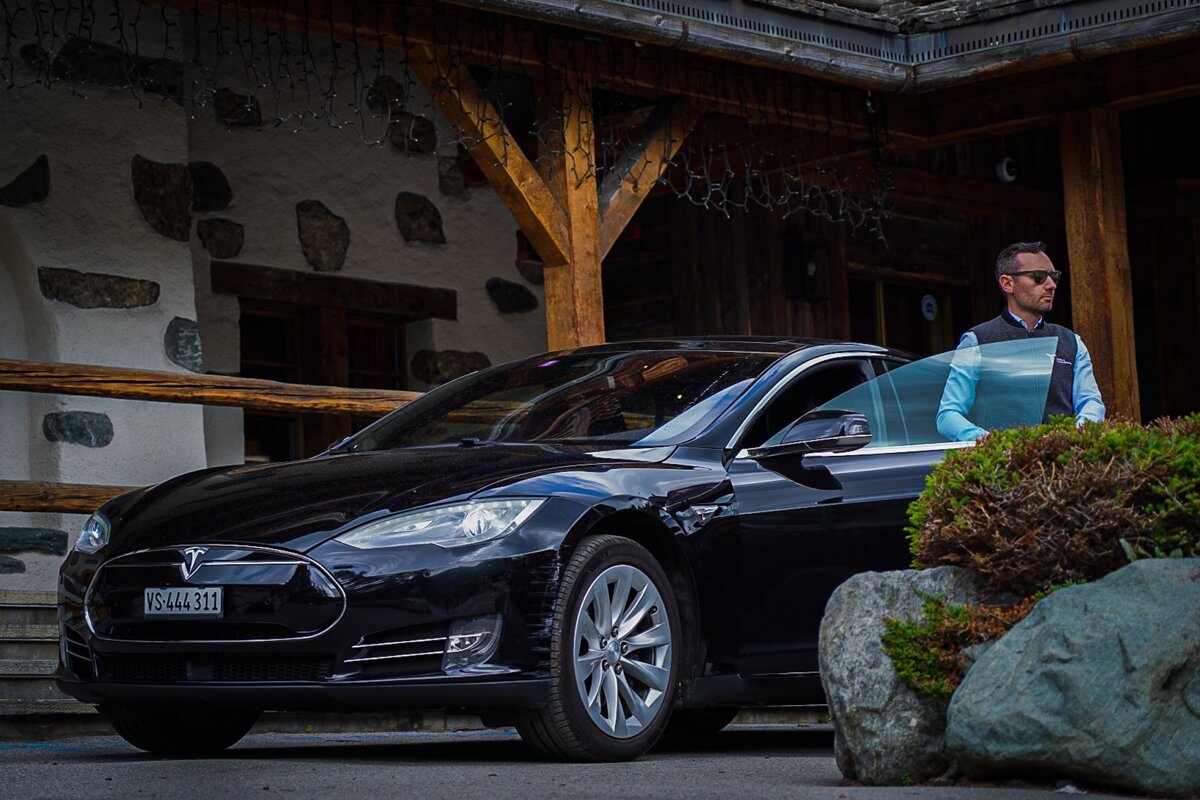 A man in sunglasses stands next to a black Tesla Model S with its door open, parked before a rustic stone and wood building.