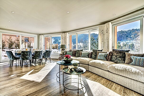 Spacious, modern living/dining room with plush seating, a dining table, and panoramic windows offering stunning snowy mountain views.