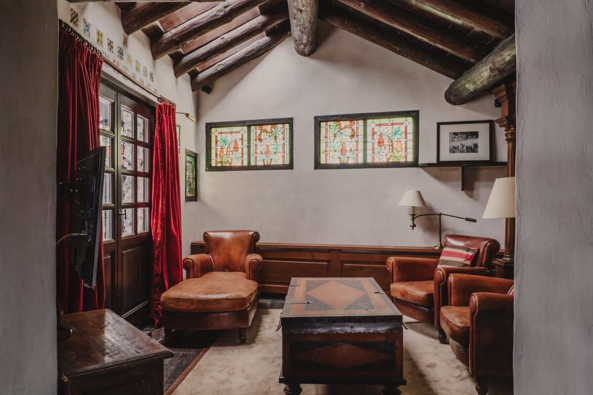 A warm, rustic living room featuring exposed wood beams, brown leather seating, red curtains, and ornate stained glass windows.
