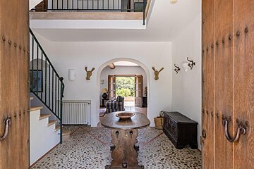 A rustic entryway with pebble flooring, open wooden doors, and archways leading to a bright outdoor area. A wooden table and staircase complete the scene.