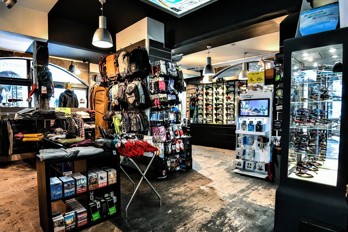 A dark retail store showcasing apparel, backpacks, and accessories on racks and shelves. Brightly lit display cases feature sunglasses, electronics, and small items.