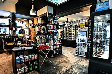 A dark retail store showcasing apparel, backpacks, and accessories on racks and shelves. Brightly lit display cases feature sunglasses, electronics, and small items.