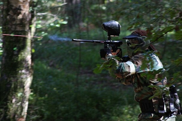 A paintball player in camouflage and mask fires a gun with a laser sight in a forest, with smoke visible from the barrel.