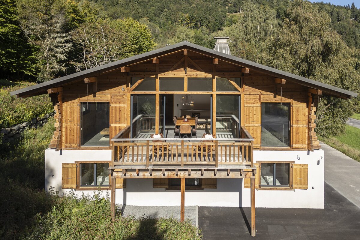 A rustic-modern wooden chalet with a large balcony and expansive windows, nestled amidst green mountains and trees.