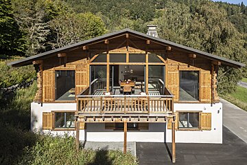 A rustic-modern wooden chalet with a large balcony and expansive windows, nestled amidst green mountains and trees.