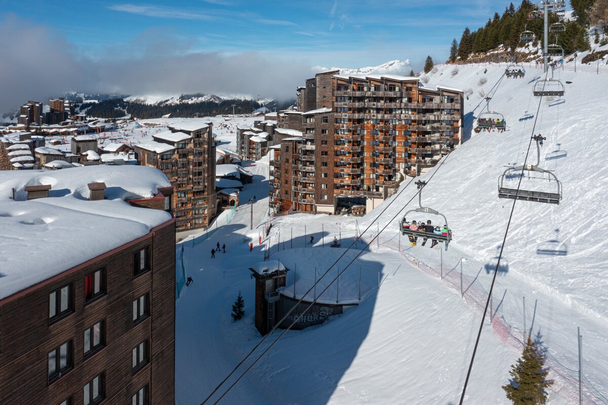 A snowy ski resort with large wooden buildings, a chairlift carrying skiers, and snow-covered slopes under a clear blue sky.