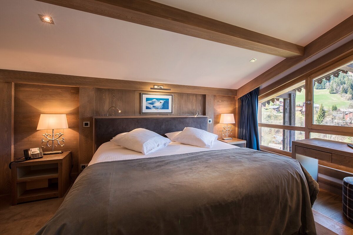 A cozy, modern chalet-style bedroom with a large bed, wooden accents, and a panoramic window offering a scenic mountain village view.